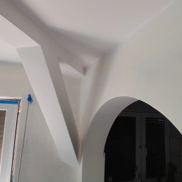Marmał-Tech - Białe, świeżo pomalowane wnętrze z łukiem i elementami konstrukcyjnymi. Okno zabezpieczone folią malarską. Minimalistyczny design, jasne oświetlenie.