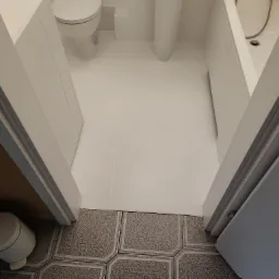 Biała łazienka po remoncie: widok z góry na nową, jednolitą posadzkę z płytek, toaletę, umywalkę i wannę. Stare płytki pod drzwiami stanowią kontrast.