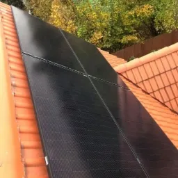 Czarne panele słoneczne zamontowane na pomarańczowej dachówce, jesienne drzewa w tle.