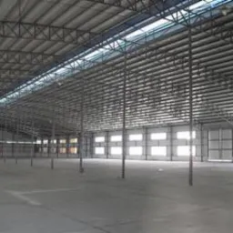 Wnętrze przestronnej hali z widoczną konstrukcją stalową dachu, oświetloną przez okna w ścianach i świetliki dachowe, surowa betonowa podłoga, puste przestrzenie.