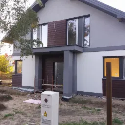 Dwukondygnacyjny dom z szarą i białą elewacją, z elementami dekoracyjnymi imitującymi drewno, widok z poziomu gruntu na niezagospodarowaną działkę.