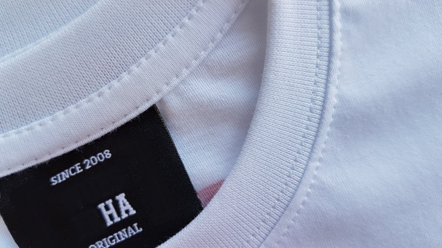 Detale wykończenia białej koszulki z bliska: widoczny szew przy dekolcie i czarna metka z napisem 'SINCE 2008 HA ORIGINAL'.