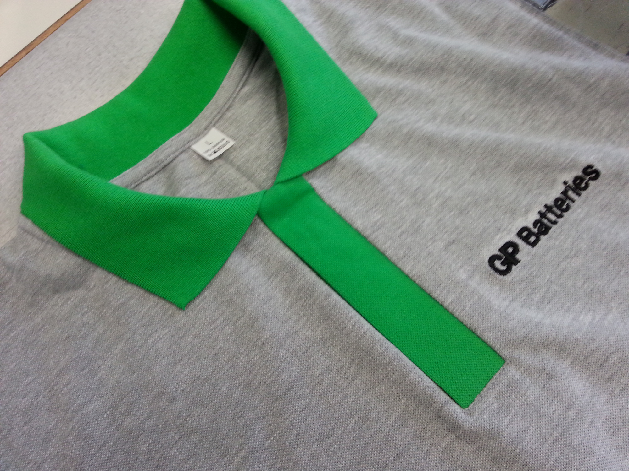 Szara koszulka polo z zielonym kołnierzykiem i logo GP Batteries, widoczna metka z rozmiarem L.