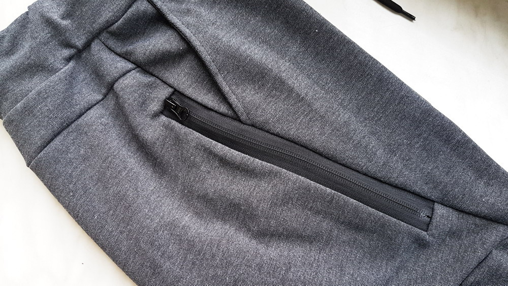 Fragment szarych dresowych spodni z wszytym czarnym zamkiem błyskawicznym przy kieszeni, widoczny sznurek