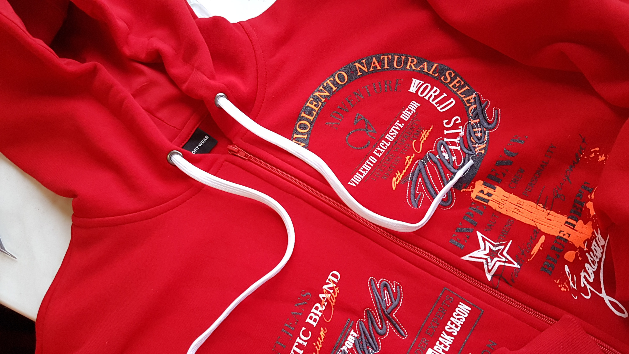 Czerwona bluza z kapturem, zapinana na zamek, z nadrukami w odcieniach szarości i pomarańczu, leżąca na jasnym tle.