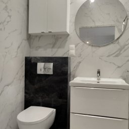 Jarosław Chróściński usługi wykończeniowe - Nowoczesna łazienka po remoncie: białe marmurowe płytki, podwieszana toaleta, szafka z umywalką i okrągłe lustro z oświetleniem LED.