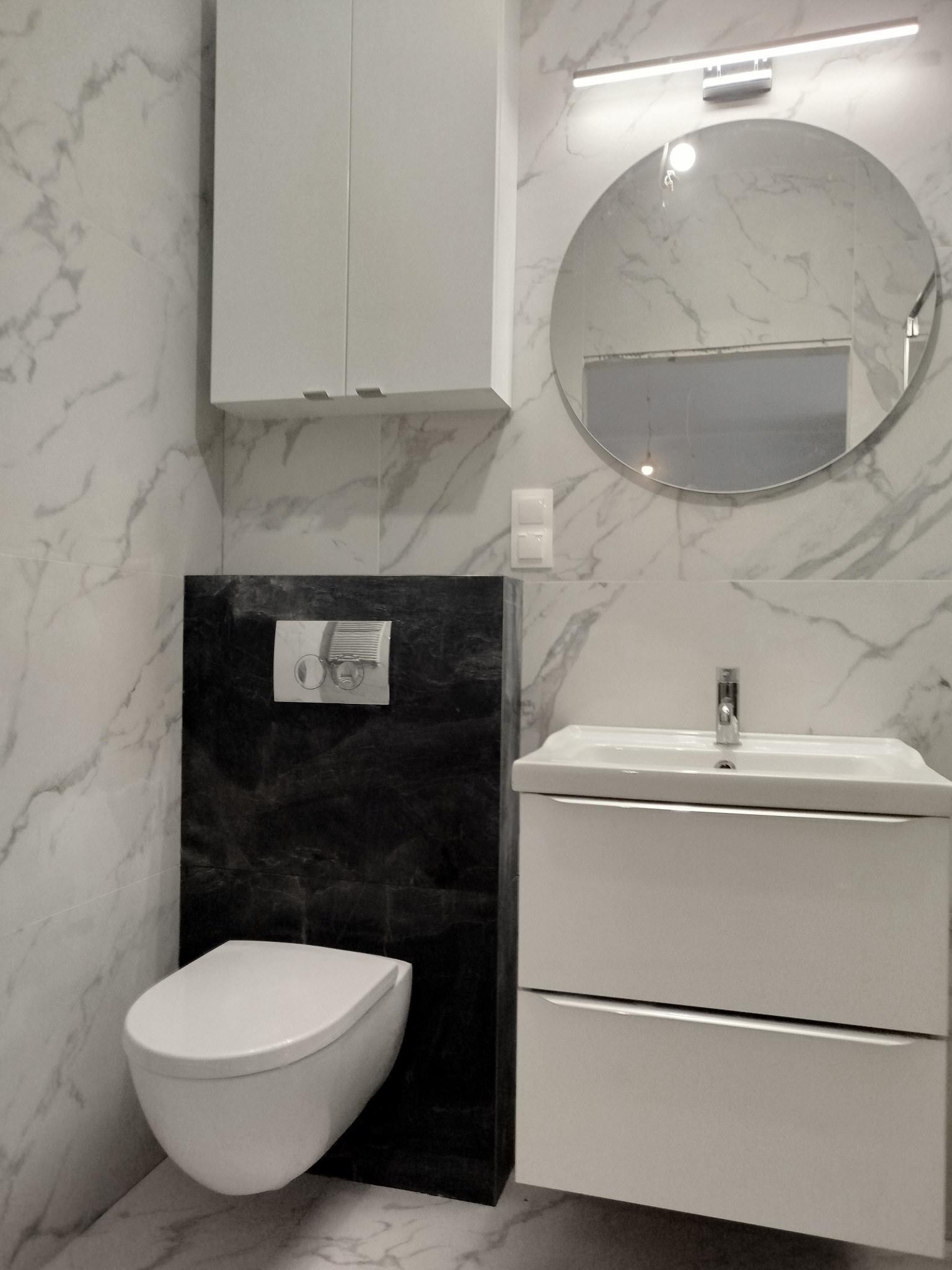 Nowoczesna łazienka po remoncie: białe marmurowe płytki, podwieszana toaleta, szafka z umywalką i okrągłe lustro z oświetleniem LED.