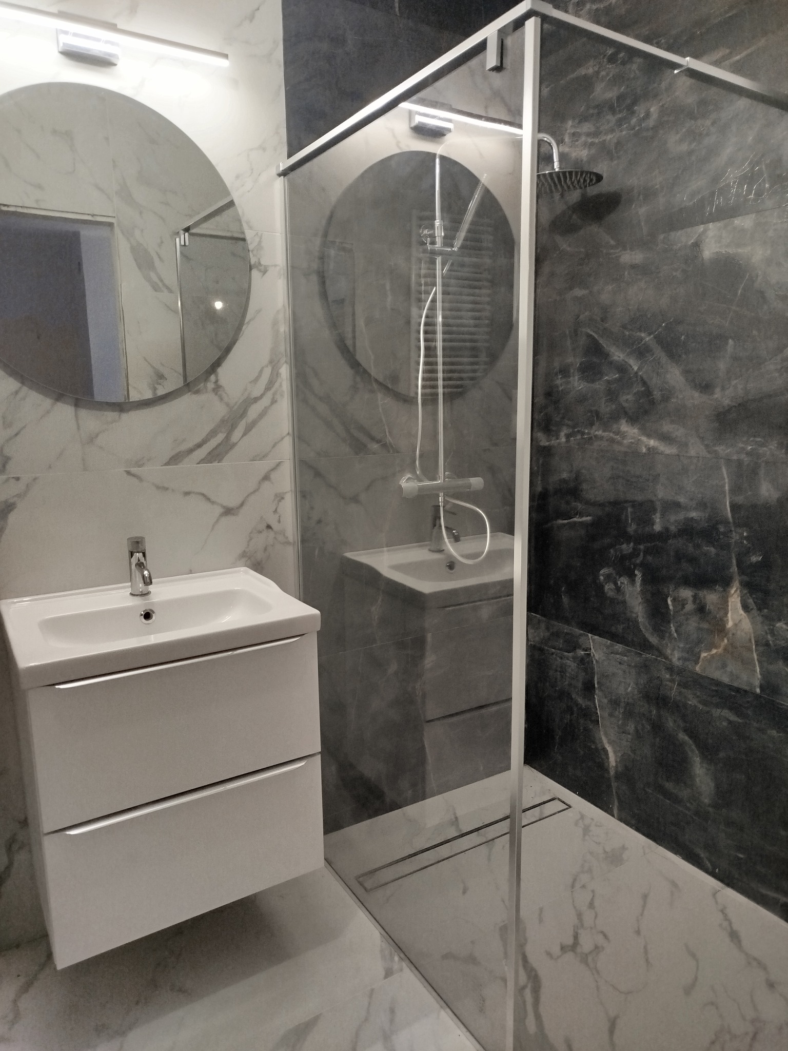Nowoczesna łazienka z kabiną prysznicową typu walk-in, umywalką z szafką i okrągłym lustrem. Marmurowe płytki w odcieniach szarości i bieli. Minimalistyczny design.