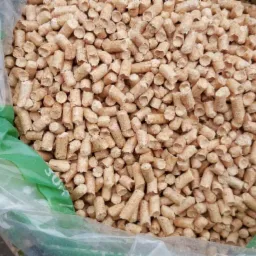 Wypełniony pelletem worek z transparentnego tworzywa, trzymany przez dłoń z czerwonym paznokciem. Widoczne drobne granulki pelletu o jasnobrązowej barwie.