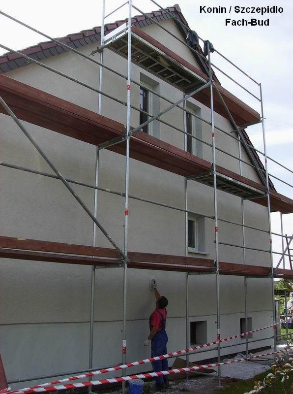 Pracownik w niebieskim kombinezonie i czerwonej koszulce wyrównuje szarą elewację domu jednorodzinnego z pomocą pacy, stojąc na rusztowaniu; w tle widoczne okna i dach budynku.