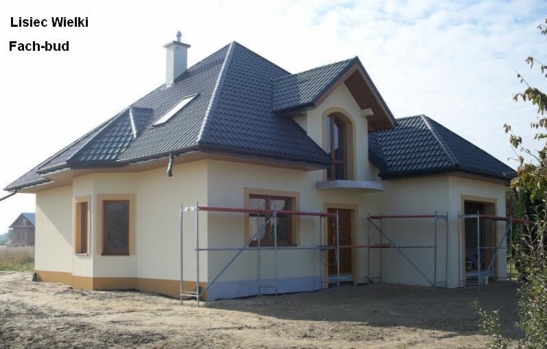 Dom jednorodzinny z kremową elewacją, ciemną dachówką i widocznymi rusztowaniami, sugerującymi trwające prace wykończeniowe.