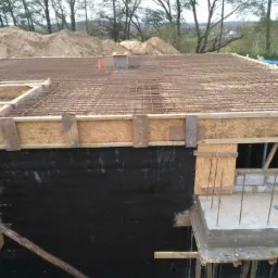 Zabezpieczone hydroizolacją fundamenty domu w trakcie budowy, zbrojenie płyty fundamentowej z widocznymi prętami stalowymi i drewnianym szalunkiem, w tle hałda piasku.
