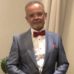 Portret mężczyzny w szarym garniturze, białej koszuli z czerwonymi guzikami i bordową muchą, stojącego na tle beżowej ściany i zasłony.