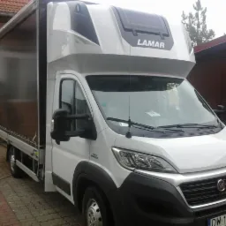 Biały bus dostawczy marki Fiat z czarną plandeką i zabudową Lamar, zaparkowany na brukowanej nawierzchni przy domu. Widoczny numer rejestracyjny.