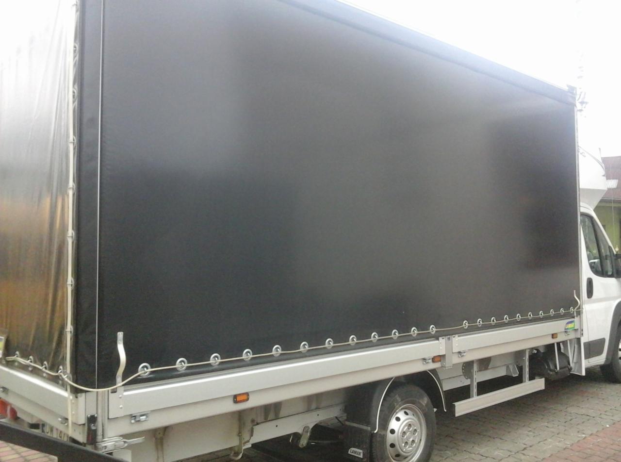 Bus z plandeką w kolorze czarnym, zaparkowany na chodniku z kostki. Widok z boku na zabudowę samochodu dostawczego.