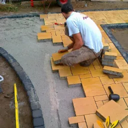 Pracownik w trakcie układania żółtej kostki brukowej na podsypce piaskowej. Widoczne narzędzia pracy, obrzeża z ciemnej kostki i taczka w tle.