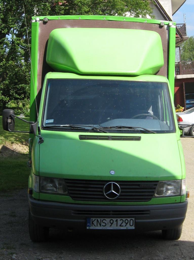Zielony Mercedes Sprinter z zabudową kontenerową, widok z przodu, tablica rejestracyjna KNS 91290, zaparkowany na trawniku przed budynkiem.