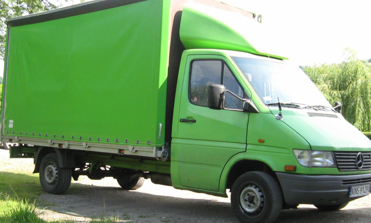 Zielony Mercedes Sprinter z plandeką na trawiastym podłożu, widoczny numer rejestracyjny KNS 91290, drzewo w tle.