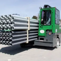 Zielony wózek widłowy z kabiną transportuje dużą ilość szarych rur PCV na palecie. Widoczne składowane rury w tle, błękitne niebo.