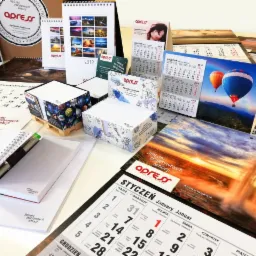 Różnorodność kalendarzy biurkowych, ściennych i notesów firmy Apress z logo i hasłem '365 dni pozytywnych emocji', prezentacja produktów na białym tle.