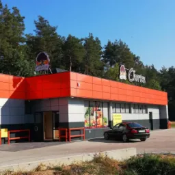 Budynek sklepu z pomarańczowo-szarą elewacją i logo sieci handlowej, widoczny parking i drzewa w tle.