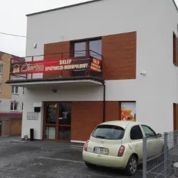 Budynek sklepu spożywczo-monopolowego z banerem reklamowym i zaparkowanym samochodem na tle miejskiego krajobrazu.