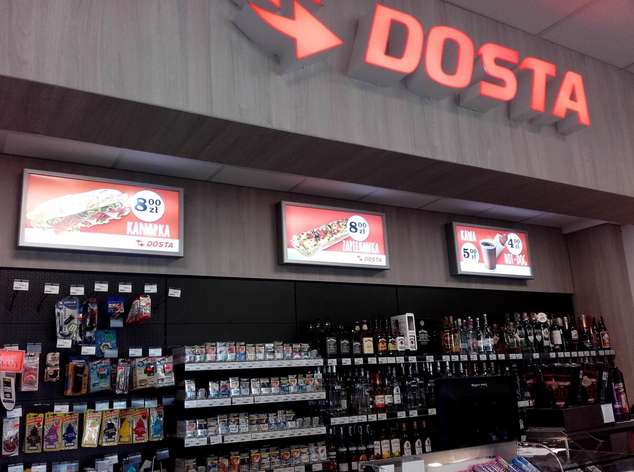 Wnętrze sklepu z podświetlanymi reklamami kanapek, zapiekanek, kawy i hot dogów, nad nimi neon 'DOSTA' ze strzałką.