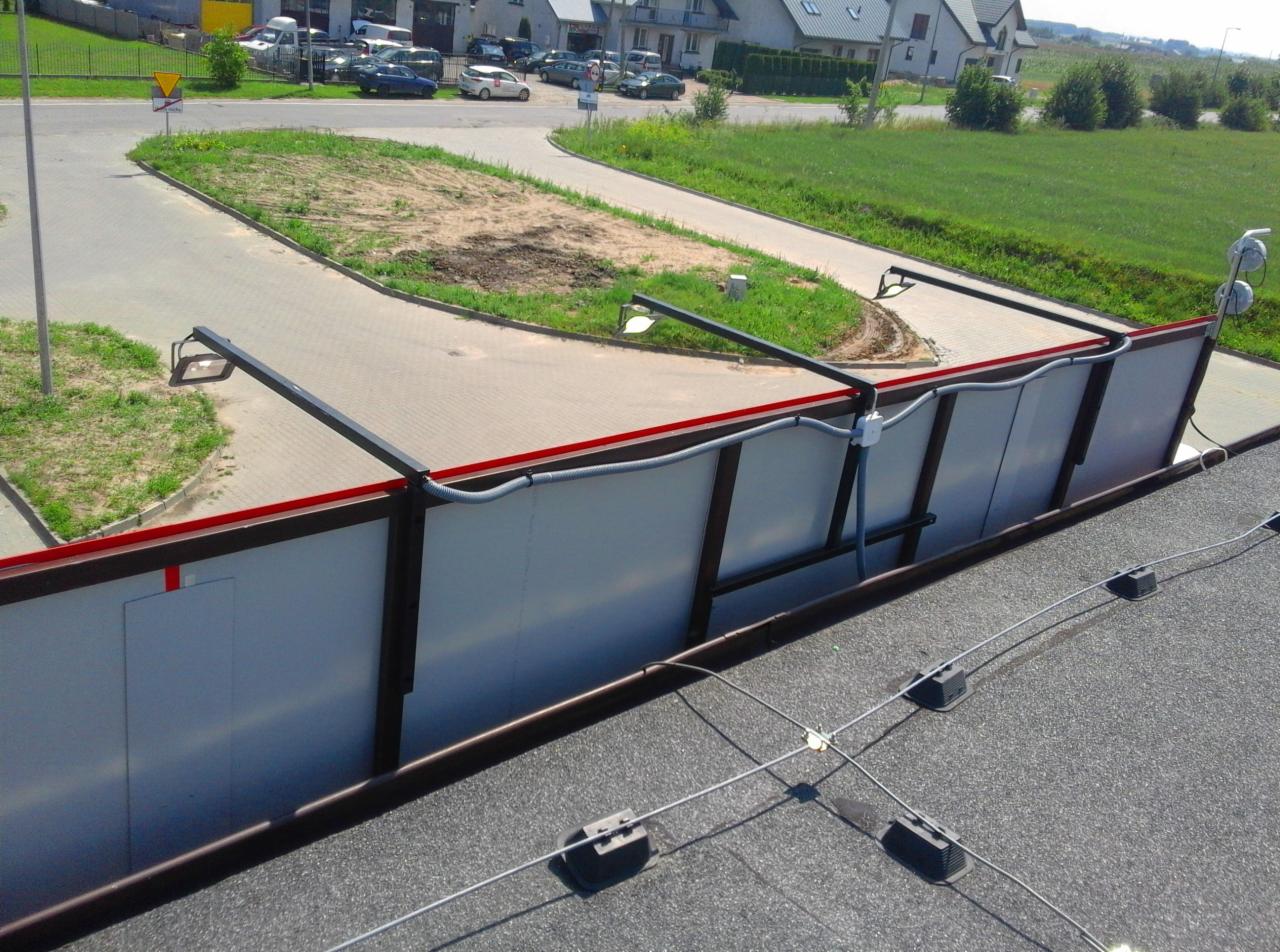 Widok z góry na konstrukcję wsporczą z oświetleniem, prawdopodobnie do reklamy wielkoformatowej, z widoczną instalacją elektryczną i fragmentem dachu budynku w tle.