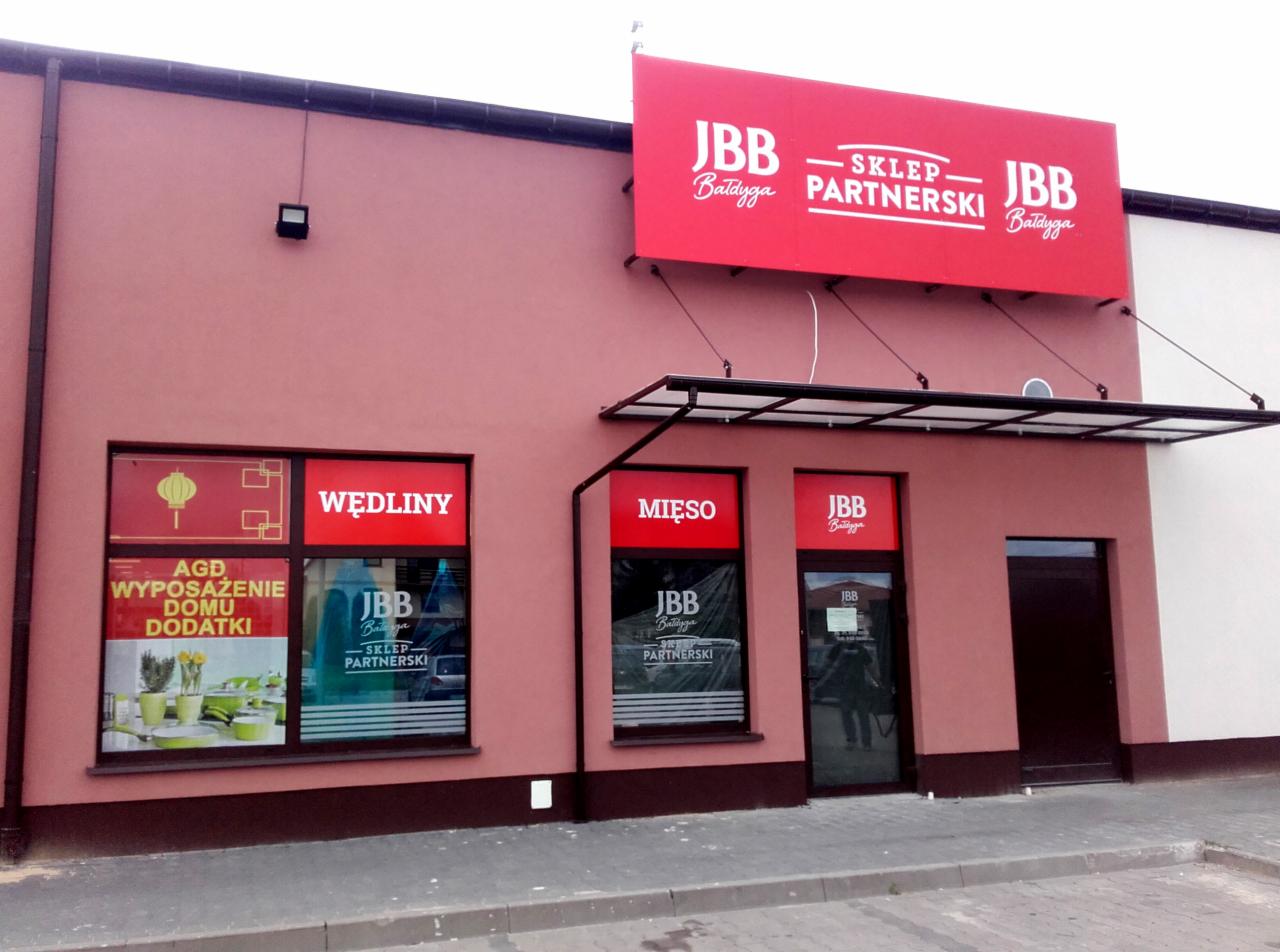 Fasada sklepu spożywczego z dużym czerwonym szyldem 'JBB Bałdyga - SKLEP PARTNERSKI JBB Bałdyga', okna z naklejkami 'Wędliny', 'Mięso' oraz 'AGD Wyposażenie Domu Dodatki'.