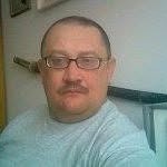 Portret mężczyzny w okularach, z wąsami, w szarej koszulce, na tle ściany z widocznym elementem dekoracyjnym. Ujęcie z bliska, oświetlenie naturalne.