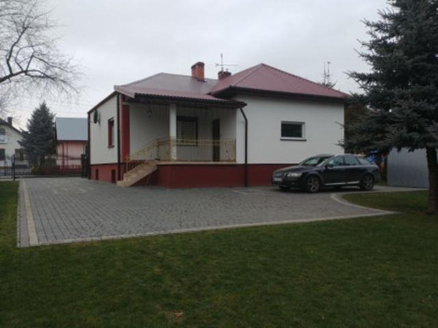 Parterowy dom z czerwonym dachem i podmurówką, szary samochód na kostce brukowej, trawnik z przodu, pochmurne niebo.