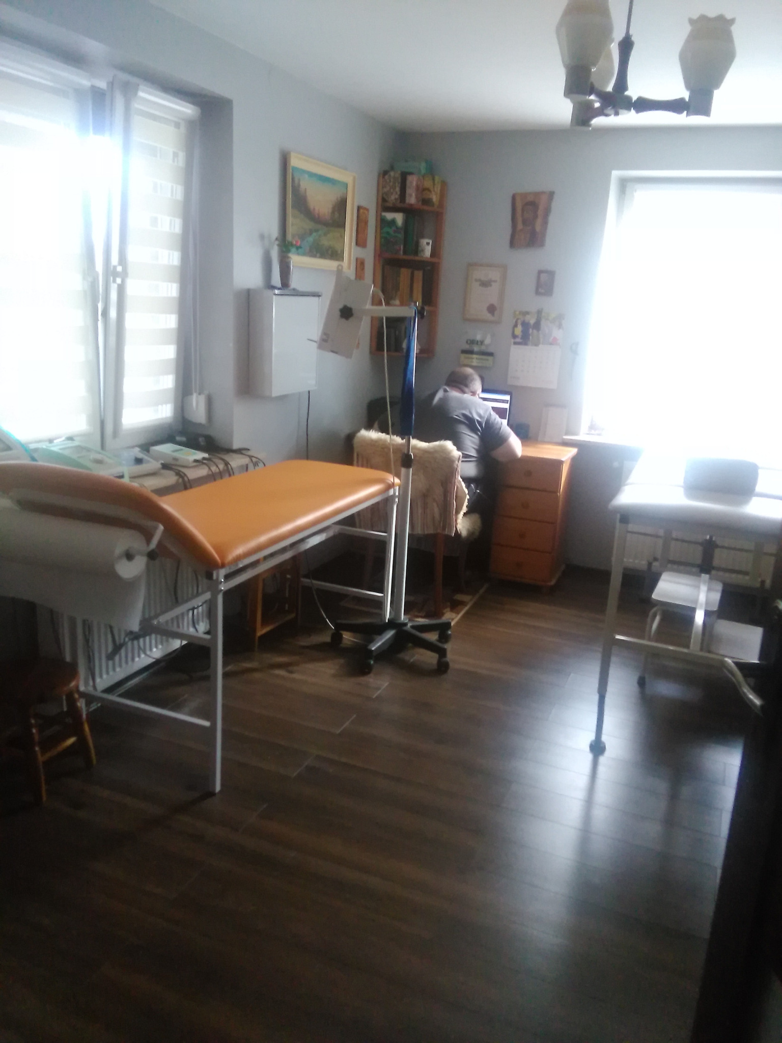 Wnętrze gabinetu z brązowymi panelami podłogowymi, kozetką fizjoterapeutyczną z pomarańczowym obiciem, biurkiem i pracującym przy nim mężczyzną, oświetlone jasnym światłem dziennym.