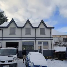 Dwukondygnacyjny dom jednorodzinny z szarą podmurówką i trzema szczytami na dachu, częściowo ośnieżony, z zaparkowanymi samochodami i osobą w kamuflażowych spodniach.