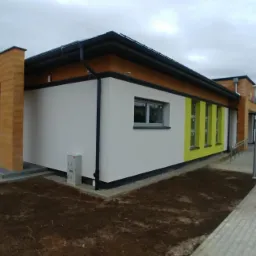 Nowoczesny budynek po termomodernizacji z białymi ścianami, drewnianymi elementami dekoracyjnymi i kolorowymi akcentami przy oknach, otoczony świeżo przygotowanym terenem.
