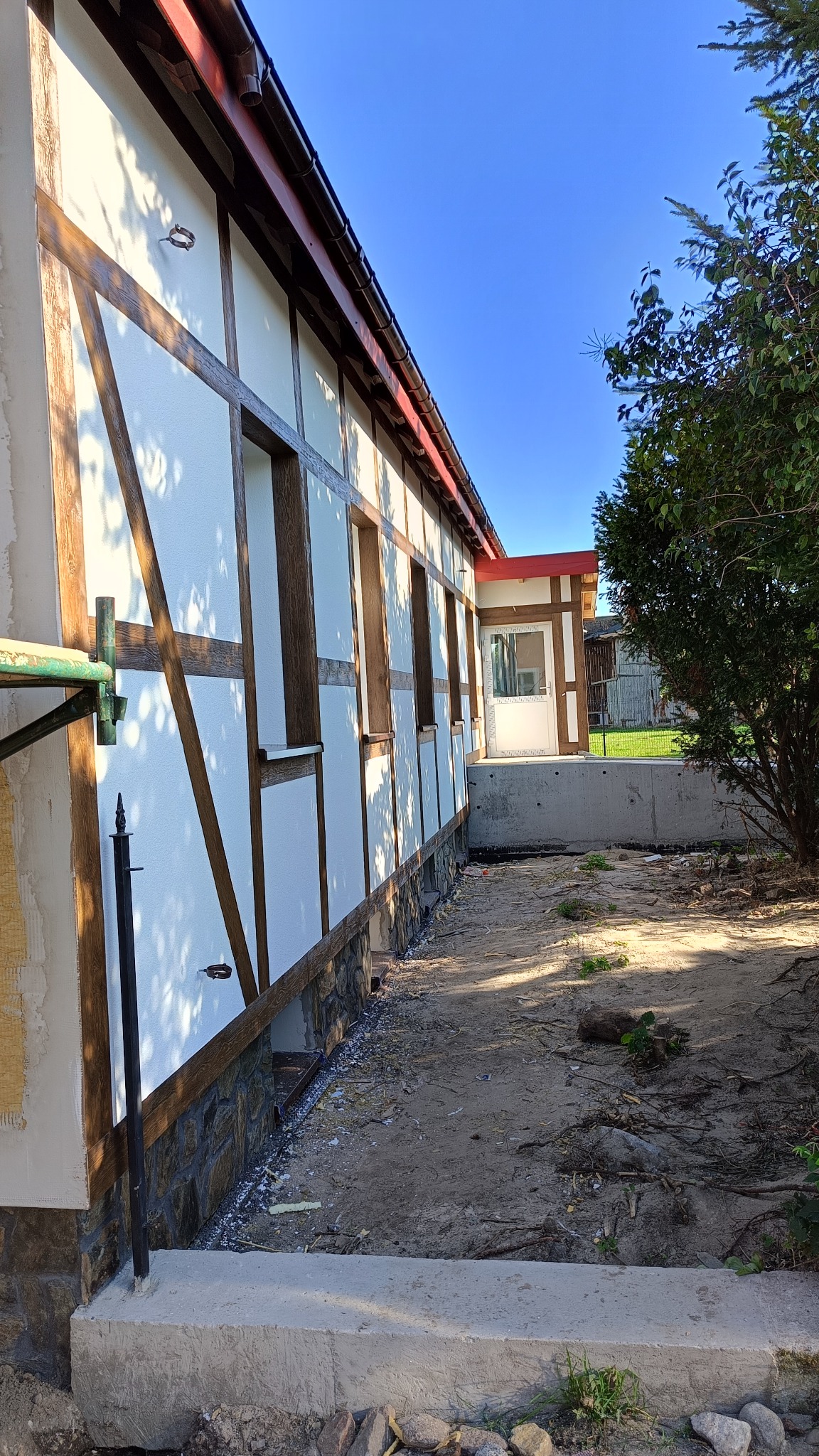 Boczny widok budynku w trakcie prac wykończeniowych elewacji, z widoczną konstrukcją szkieletową z drewnopodobnych belek, ocieplonymi panelami i podmurówką z kamienia naturalnego, na tle błękitnego...