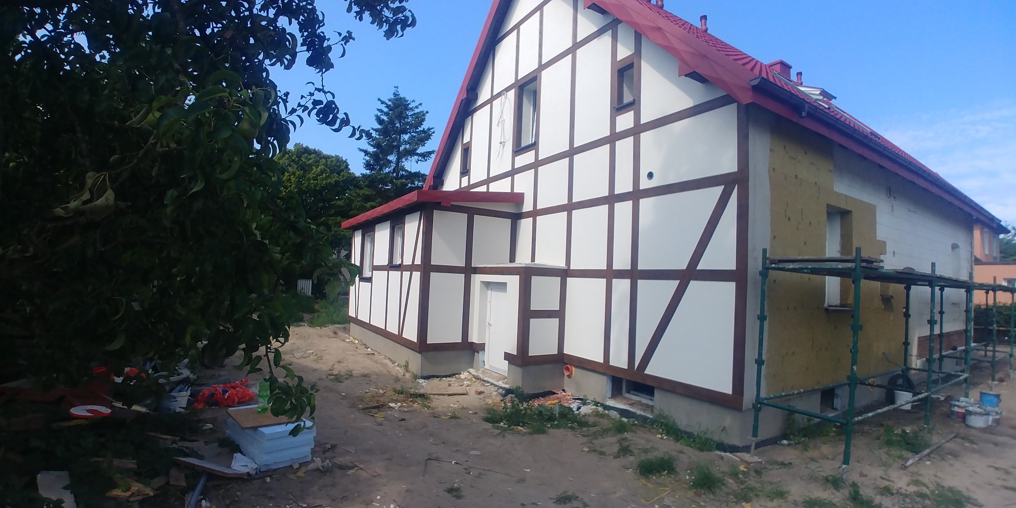 Dom w trakcie prac elewacyjnych: część ściany ocieplona wełną mineralną, druga część wykończona w stylu szachulcowym, widoczne rusztowania i materiały budowlane.