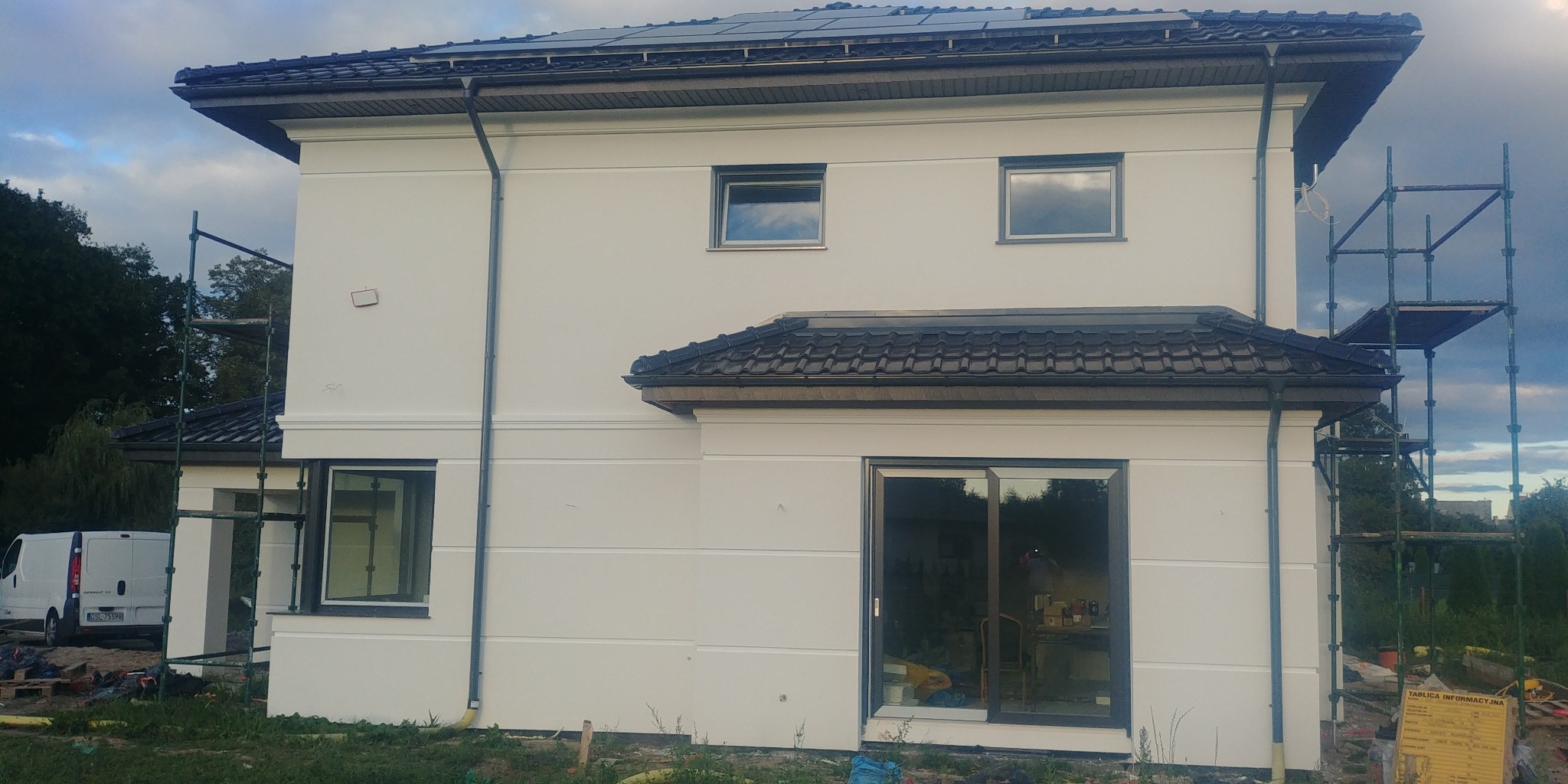 Dwupiętrowy dom z białą elewacją, widoczne rusztowania i zaparkowany biały bus, sugerujące prace wykończeniowe na zewnątrz budynku.