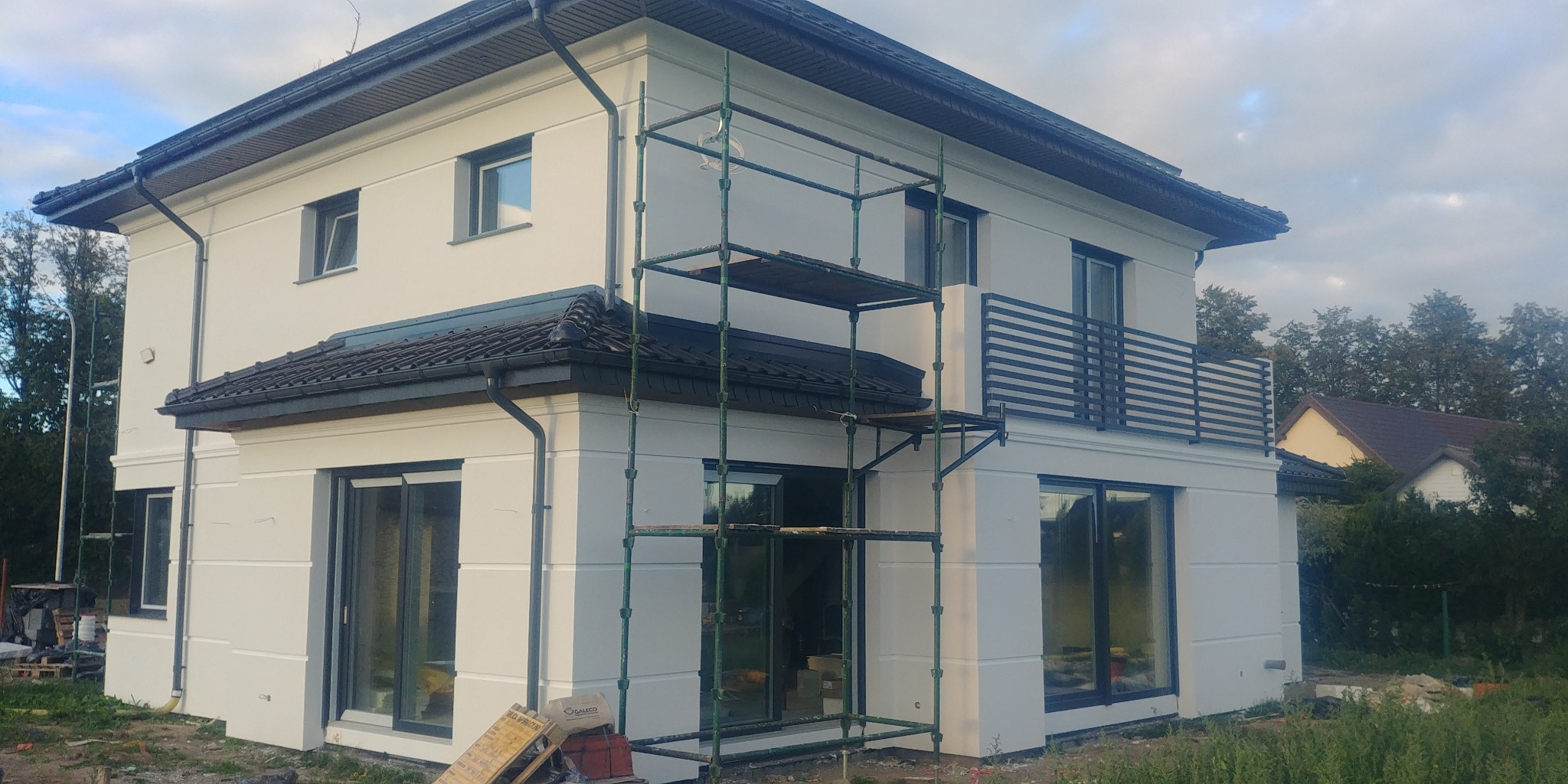 Dwukondygnacyjny dom jednorodzinny z jasną elewacją, widoczne rusztowanie przy ścianie, balkon z czarną balustradą i ciemna dachówka.