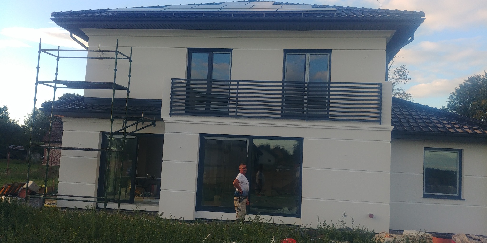 Nowoczesny dom z ocieploną białą elewacją, balkonem z czarną balustradą i panelami słonecznymi na dachu, widoczny fragment rusztowania i pracownik w tle.