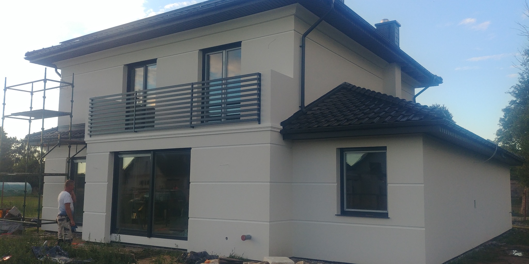 Dwukondygnacyjny dom jednorodzinny z jasną elewacją, czarną balustradą balkonową i fragmentem rusztowania po lewej stronie, widoczny pracownik w tle.