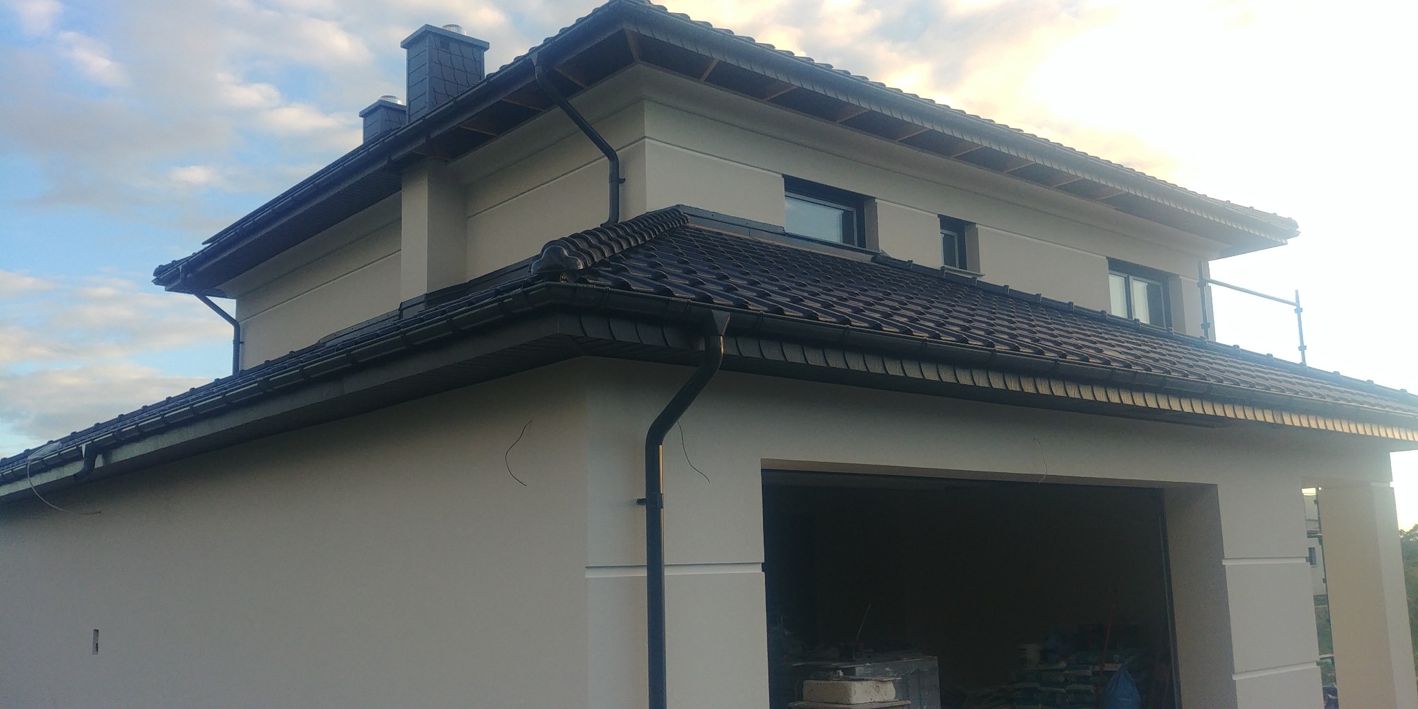 Jasny, dwupiętrowy dom z garażem i ciemną dachówką, widoczny z boku, z elementami dekoracyjnymi na elewacji i rurami spustowymi.