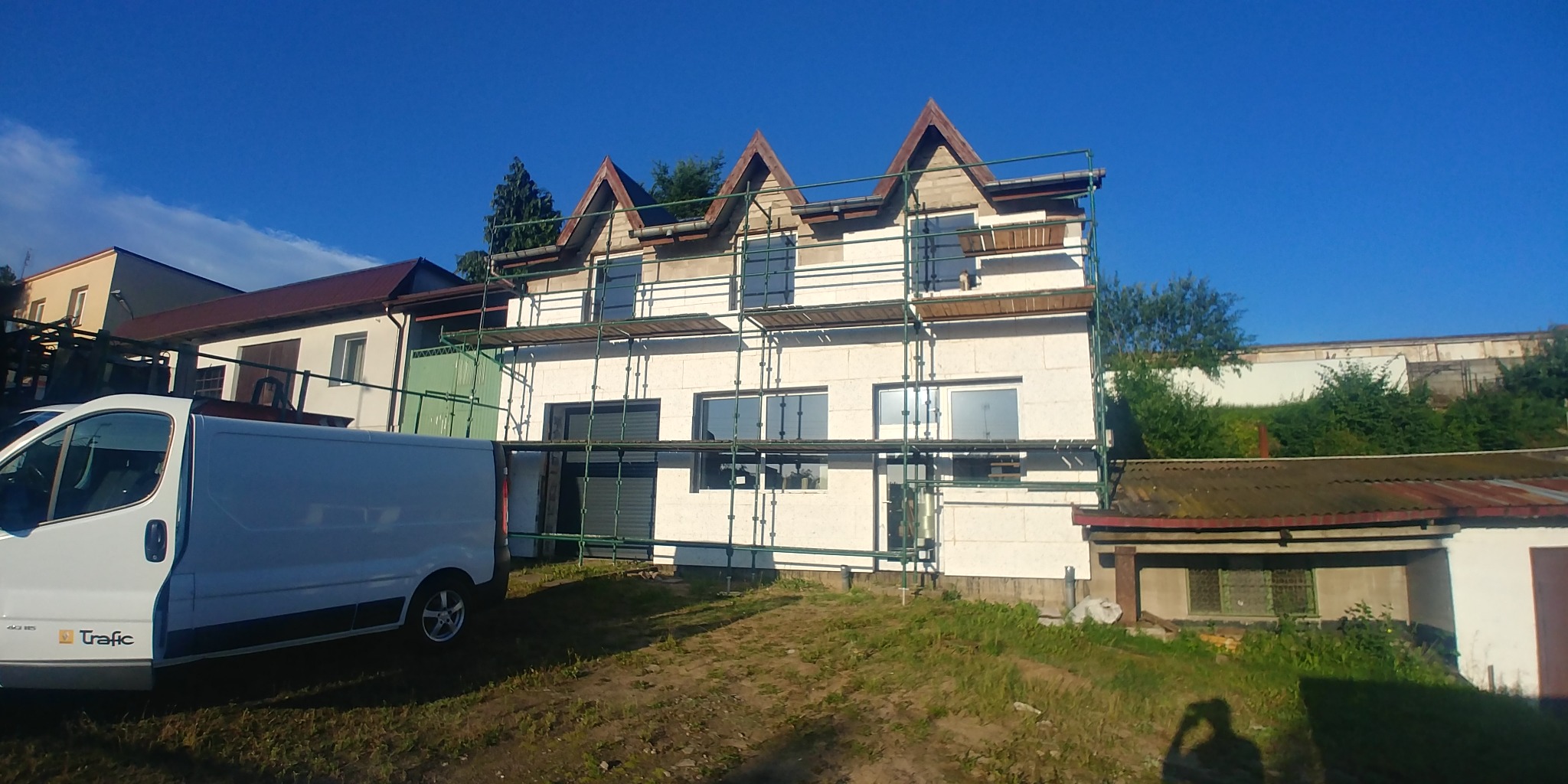 Dwupiętrowy dom w trakcie prac elewacyjnych, z widocznymi rusztowaniami, częściowo ocieploną ścianą i zaparkowanym białym vanem z otwartymi drzwiami bocznymi na pierwszym planie.