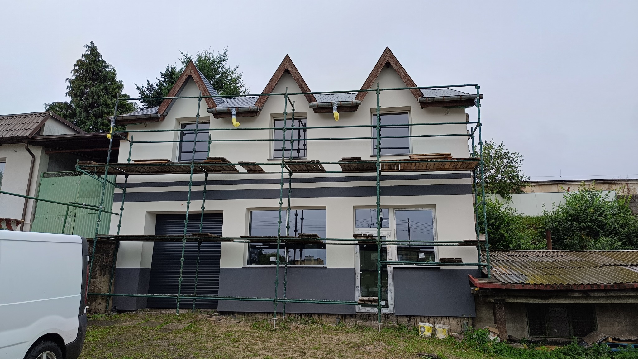 Dom jednorodzinny w trakcie prac wykończeniowych elewacji, widoczne rusztowania i częściowo pomalowana fasada w kolorze białym i szarym, trzy szczyty z drewnianym wykończeniem, pochmurne niebo w tle.