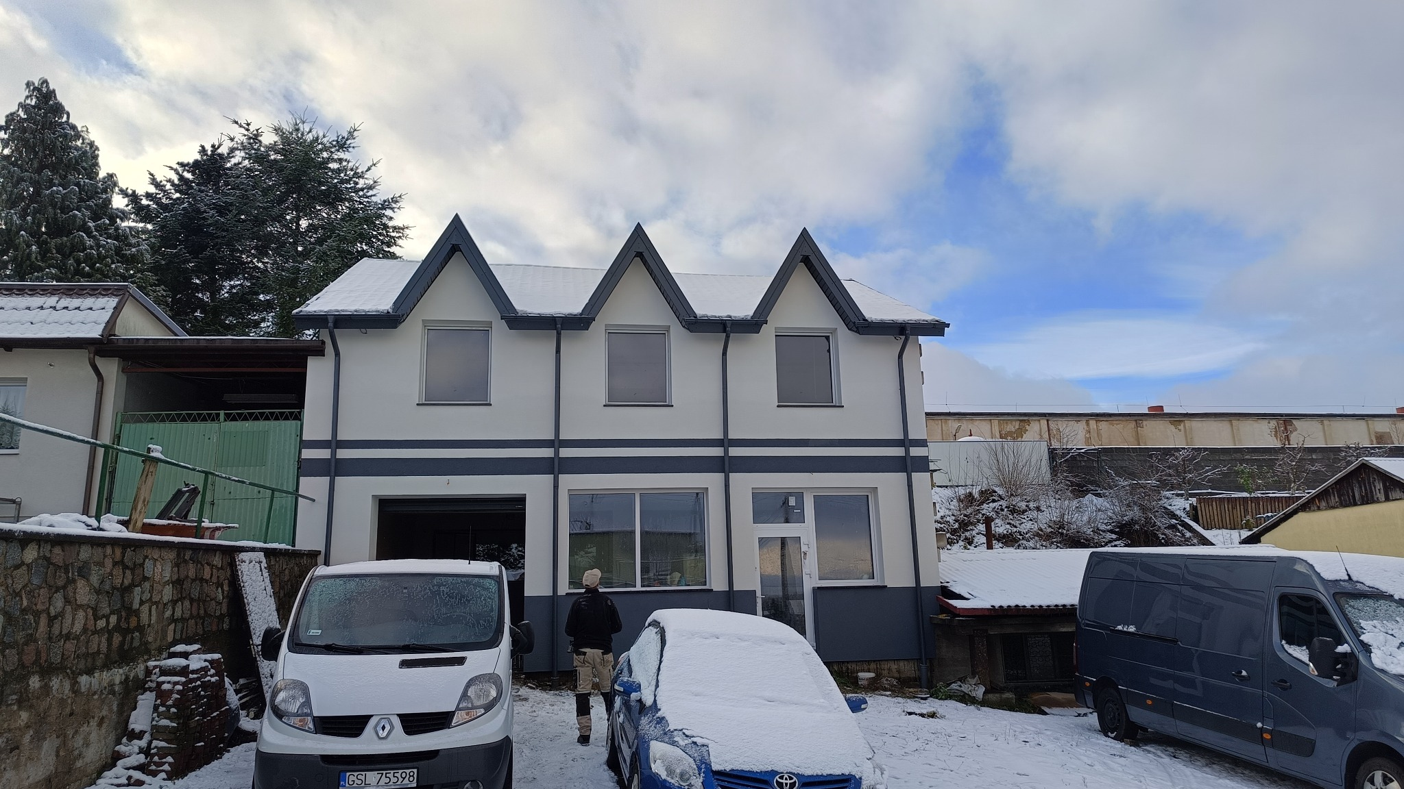 Dwukondygnacyjny dom jednorodzinny z szarą podmurówką i trzema szczytami na dachu, częściowo ośnieżony, z zaparkowanymi samochodami i osobą w kamuflażowych spodniach.