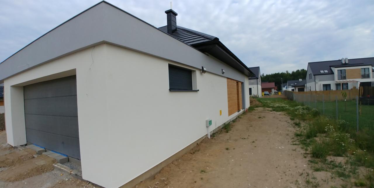 Nowoczesny dom jednorodzinny z jasną elewacją, grafitowym dachem i garażem, widok z boku na tle pochmurnego nieba i innych domów w zabudowie szeregowej.