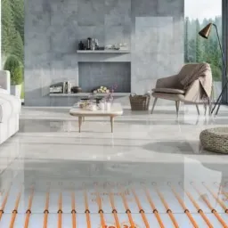 Salon z ogrzewaniem podłogowym: widoczne pomarańczowe kable grzewcze pod błyszczącą, jasną posadzką, w tle sofa, fotel i dekoracyjny kominek.