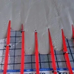 Wylewka anhydrytowa w trakcie rozpływania się wokół czerwonych rurek ogrzewania podłogowego, widoczne metalowe klamry mocujące rurki do podłoża.