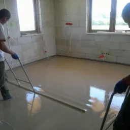 Wylewanie posadzki w pomieszczeniu z oknami. Dw&oacute;ch mężczyzn rozprowadza masę poziomującą. Widoczne ściany z pustak&oacute;w i instalacje elektryczne.