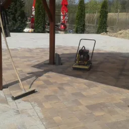 Ułożona kostka brukowa w odcieniach brązu i beżu pod drewnianą konstrukcją, z ubijarką i szczotką. Częściowo zasypana piaskiem.