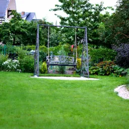 Metalowa huśtawka ogrodowa z ozdobnym siedziskiem, otoczona zadbanym trawnikiem i r&oacute;żnorodną roślinnością, w tle dom jednorodzinny.